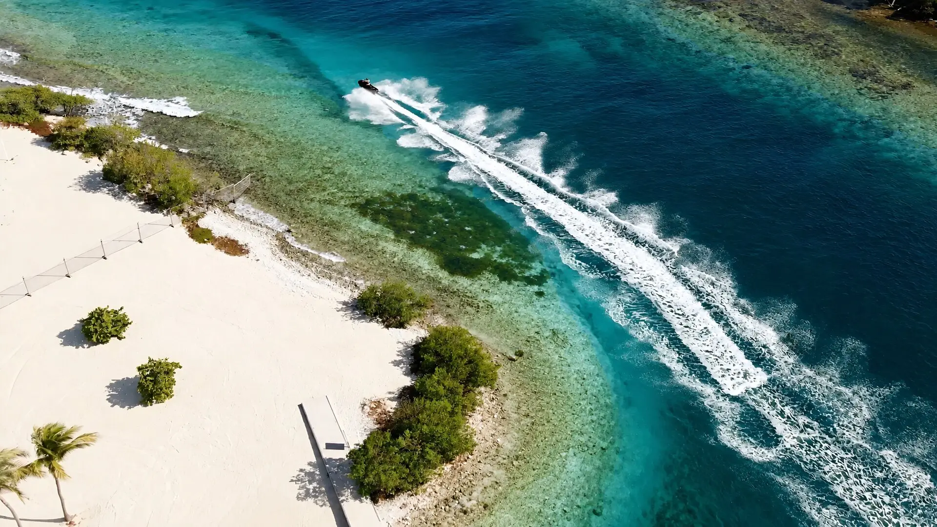 Le sillage d'un jet ski longe une plage de sable blanc, entre l'eau turquoise et le bleu du large, vu du ciel