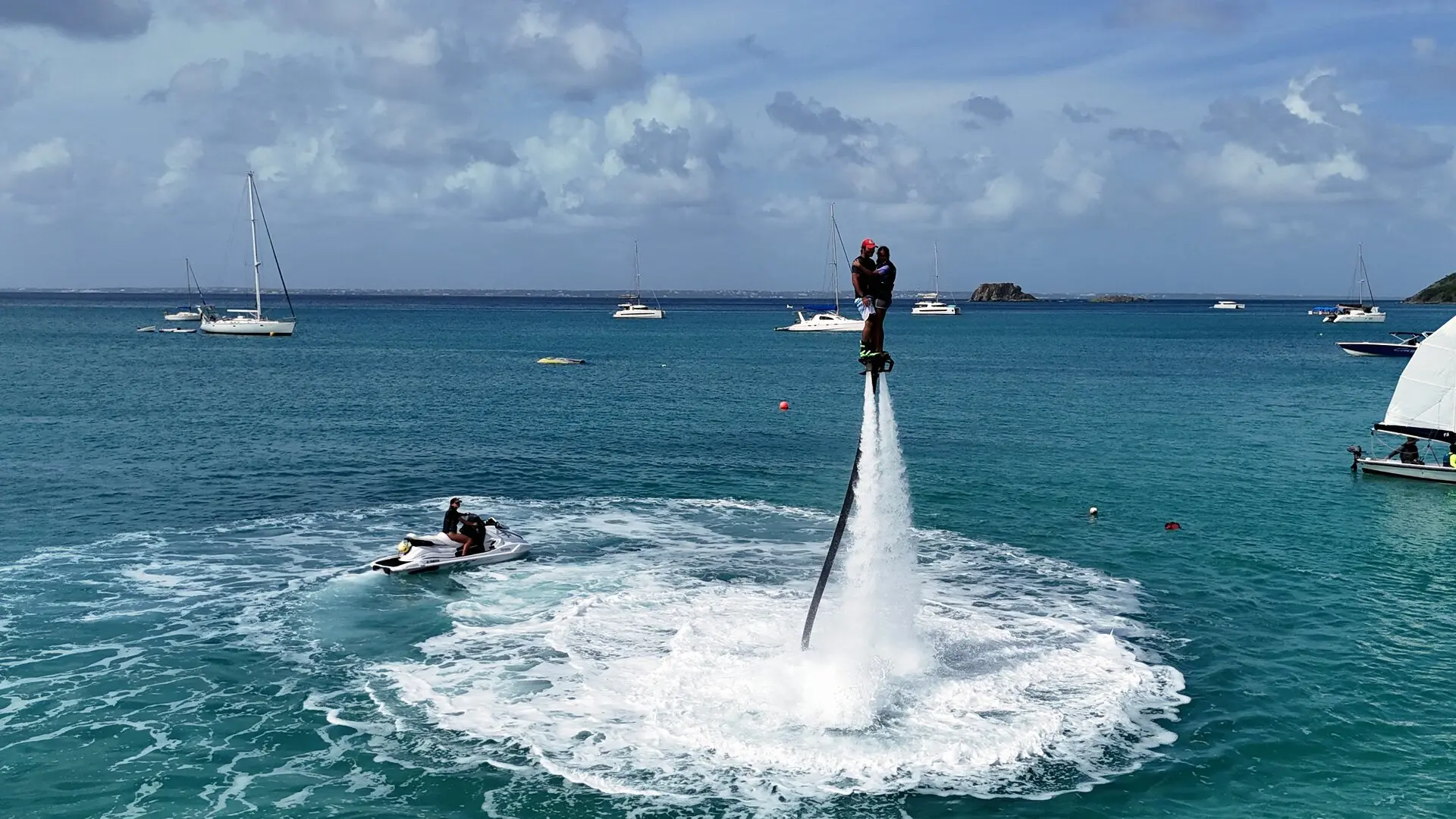 Deux personnes en flyboard au-dessus de l'eau, un jet ski à côté, des voiliers au mouillage